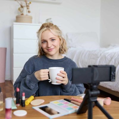 Portrait of beautiful social media beauty blogger, sitting in front of digital camera on floor in bedroom, drinking tea and chatting, talking to followers.